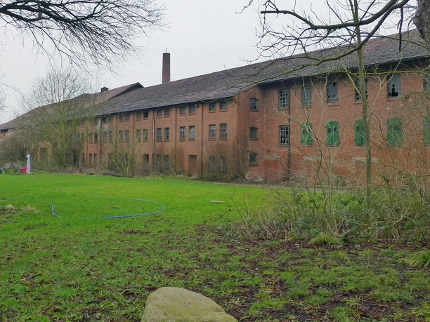 Altes Backsteingebäude mit grünen Fensterläden, umgeben von grüner Wiese und kahlen Bäumen.