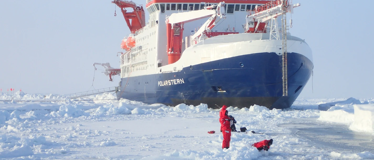 Forschende in roten Anzügen nehmen Eisproben vor der Polarstern im arktischen Packeis.