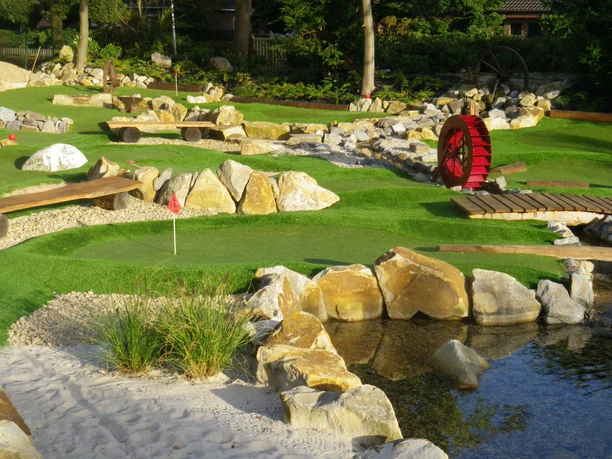 Abenteuergolf Janßen Hof Abenteuergolf-Anlage mit Felsen, Wasserlauf, Holzbrücken und rotem Wasserrad.