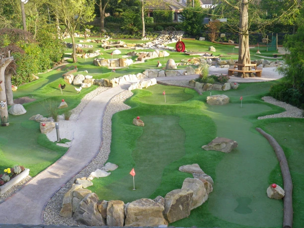 Abenteuergolf Janßen Hof Abenteuergolfanlage mit Kunstrasenbahnen, Felsen, roten Fahnen und Holztisch unter Bäumen.