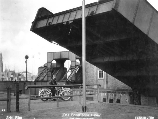 Schwarzweißszene: geöffnete Klappbrücke über einem Mann mit Fahrrad am Hafen.