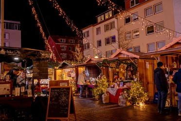 Beleuchtete Holzhütten, Tannen und Besucher auf einem Weihnachtsmarkt bei Nacht.