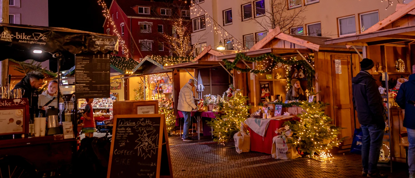 Weihnachtsmarkt_mit_Familie_2025-24__c__Carmen_Lindemann_Erlebnis_Bremerhaven_CC0_WEB.jpg Beleuchtete Holzhütten, Tannen und Besucher auf einem Weihnachtsmarkt bei Nacht.