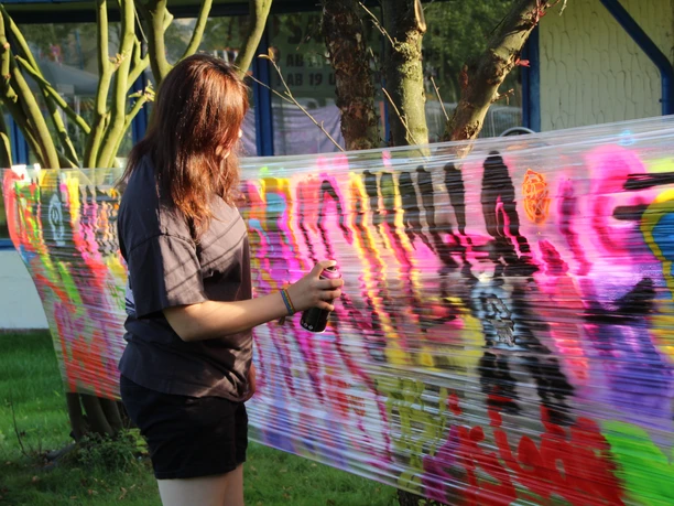 Junge Person sprüht buntes Graffiti auf gespannte Folie zwischen Bäumen.