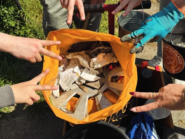 Hände zeigen auf gesammelten Müll in orangefarbener Tüte bei einer Clean-up-Aktion.
