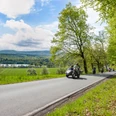 Eine Landstraße führt am Ufer eines Sees entlang, gesäumt von grünen Bäumen, während Motorräder passieren.