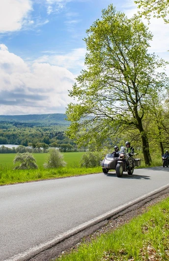 Eine Landstraße führt am Ufer eines Sees entlang, gesäumt von grünen Bäumen, während Motorräder passieren.