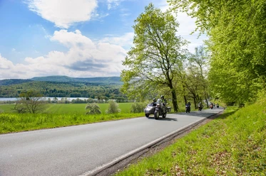 Schieder-See Eine Landstraße führt am Ufer eines Sees entlang, gesäumt von grünen Bäumen, während Motorräder passieren.
