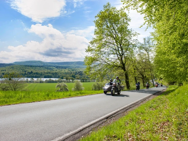 Schieder-See Eine Landstraße führt am Ufer eines Sees entlang, gesäumt von grünen Bäumen, während Motorräder passieren.