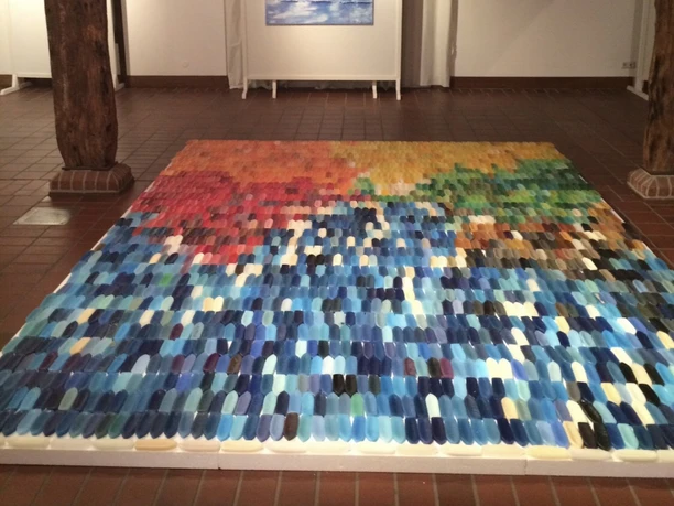 Bunte Wachsschiffchen-Installation in einer Ausstellung, dahinter ein Meeresbild.