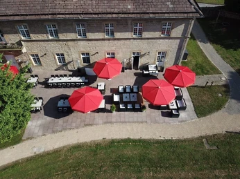 Luftaufnahme einer Terrasse mit roten Sonnenschirmen neben einem historischen Gebäude.