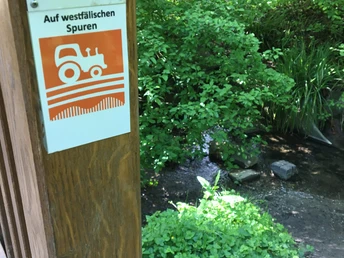 Schild mit Traktor-Piktogramm und Aufschrift „Auf westfälischen Spuren“ vor üppigem Garten mit Bach.