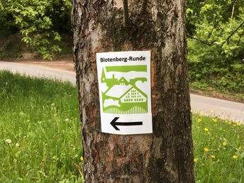 Markierungszeichen am Wegesrand Wegweiser für die "Blotenberg-Runde" an einem Baum in einer grünen Landschaft mit Pfeilrichtung.