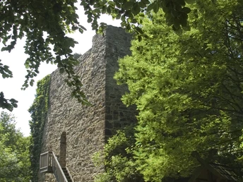 Eine alte Steinruine mit Außentreppe umgeben von üppigem Grün, Menschen spazieren und sitzen nahebei.