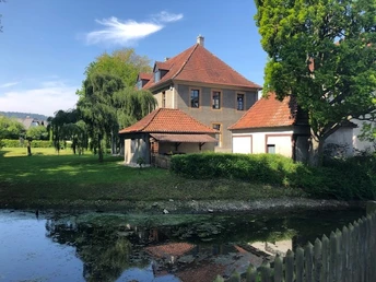Bürgerbegegnungsstätte Haus Werther Historisches Gebäude mit roten Ziegeldächern am Ufer eines Wassergrabens, umgeben von Bäumen.
