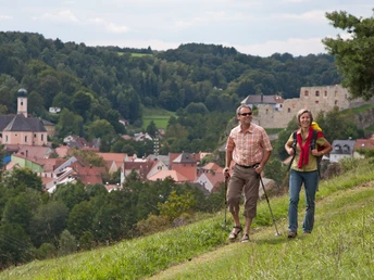 Am Burgensteig Nähe Laaber Zwei Wanderer auf einem grünen Hügel überblicken ein Dorf mit einer Kirche und einer Burgruine.