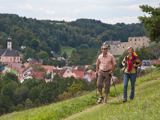 Am Burgensteig Nähe Laaber Zwei Wanderer auf einem grünen Hügel überblicken ein Dorf mit einer Kirche und einer Burgruine.