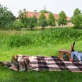 Picknickdecke mit Korb und Schuhen auf ländlicher Wiese, im Hintergrund Bäume und ein Gebäude.