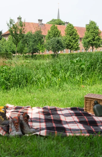 Picknickdecke mit Korb und Schuhen auf ländlicher Wiese, im Hintergrund Bäume und ein Gebäude.