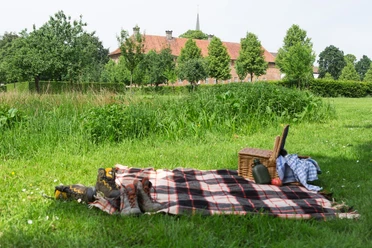 Das Kloster Clarholz beim Picknick im Blick Picknickdecke mit Korb und Schuhen auf ländlicher Wiese, im Hintergrund Bäume und ein Gebäude.
