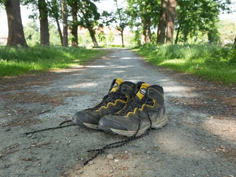 Ein Paar abgenutzte Wanderstiefel liegt auf einem Schotterweg, umgeben von hohen, grünen Bäumen.