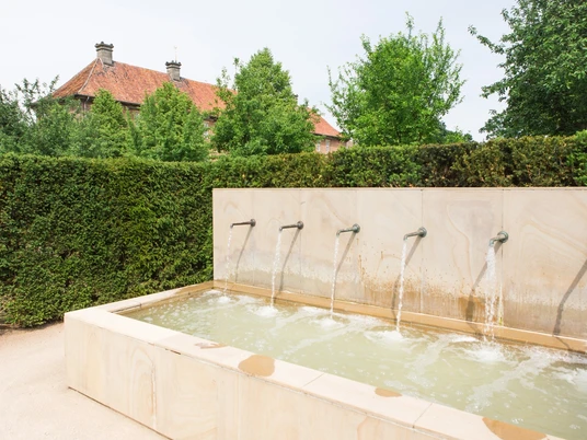 Ein moderner Brunnen mit sechs Wasserausläufen vor einer Hecke und einem roten Ziegelgebäude.