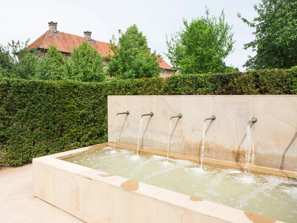 Ein moderner Brunnen mit sechs Wasserausläufen vor einer Hecke und einem roten Ziegelgebäude.