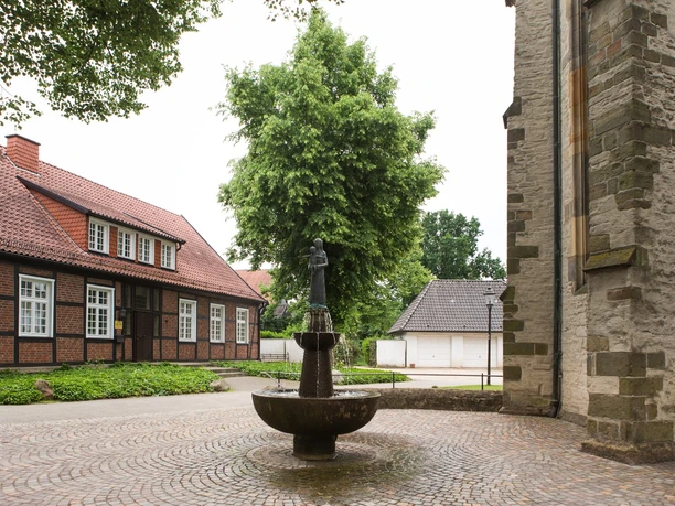 Kirchplatz mit Brunnen Ein historischer Brunnen mit Figur vor Fachwerk- und Steinhäusern unter einem bewölkten Himmel.