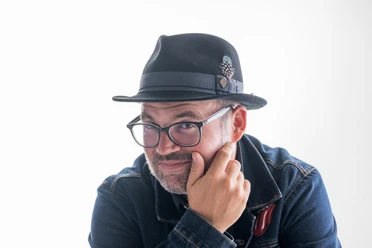 Björn Heuser Porträt von Björn Heuser mit Hut, Brille und Jeansjacke vor hellem Hintergrund