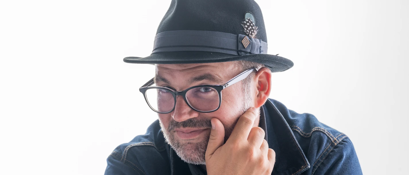 Björn Heuser Porträt von Björn Heuser mit Hut, Brille und Jeansjacke vor hellem Hintergrund