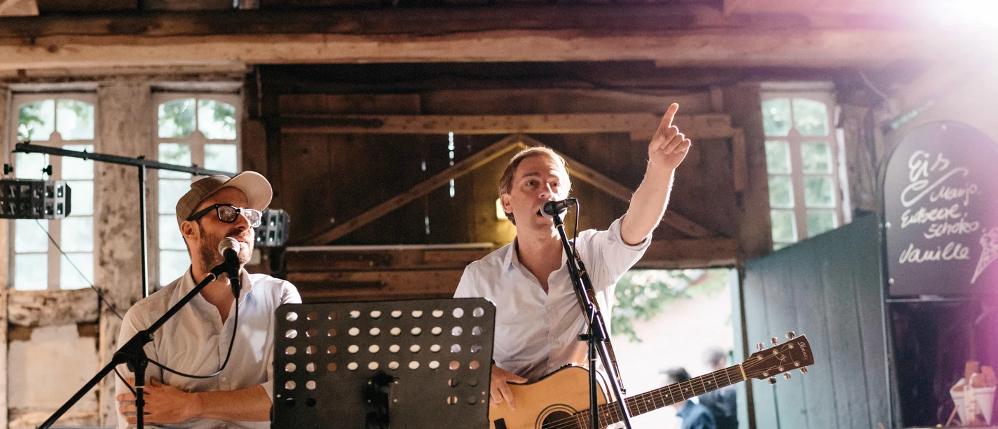 Songbingo-twinns Zwei Musiker mit Mikrofonen und Gitarre bei einem Songbingo-Auftritt.