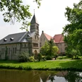 Historische Kirche mit steinernen Details und Turm, umgeben von grünen Bäumen, Teich im Vordergrund.