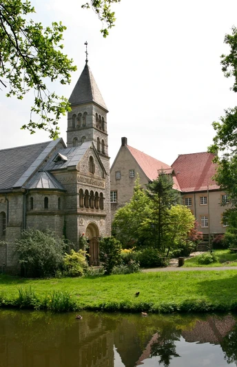 Historische Kirche mit steinernen Details und Turm, umgeben von grünen Bäumen, Teich im Vordergrund.