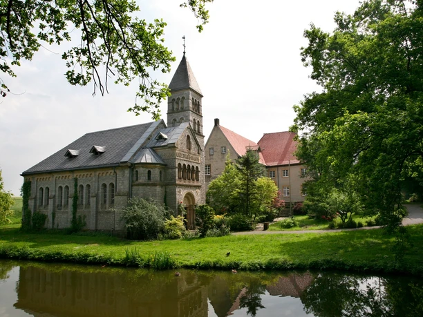 Historische Kirche mit steinernen Details und Turm, umgeben von grünen Bäumen, Teich im Vordergrund.