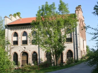 Historische Backsteinfabrik mit roten Ziegeldach, umgeben von Bäumen und einem blauen Himmel.