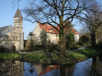 Historisches Kloster mit romanischer Kirche, Baum und einem kleinen Wassergraben im Vordergrund.