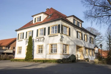 Zweistöckiges weißes Gebäude mit roten Ziegeldach und grünen Fensterläden an einem sonnigen Tag.
