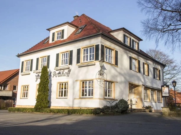 Zweistöckiges weißes Gebäude mit roten Ziegeldach und grünen Fensterläden an einem sonnigen Tag.