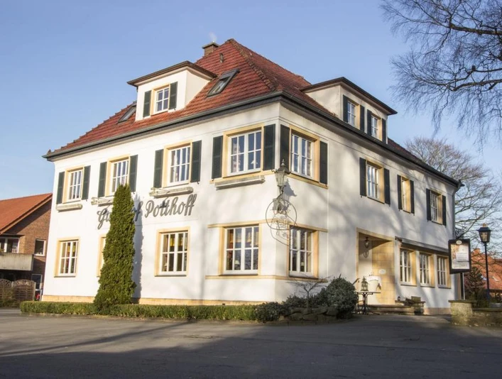 Zweistöckiges weißes Gebäude mit roten Ziegeldach und grünen Fensterläden an einem sonnigen Tag.