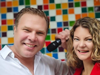 Schmitzebud Duo A smiling Schmitzebud duo with a microphone in front of a colorful tiled wall