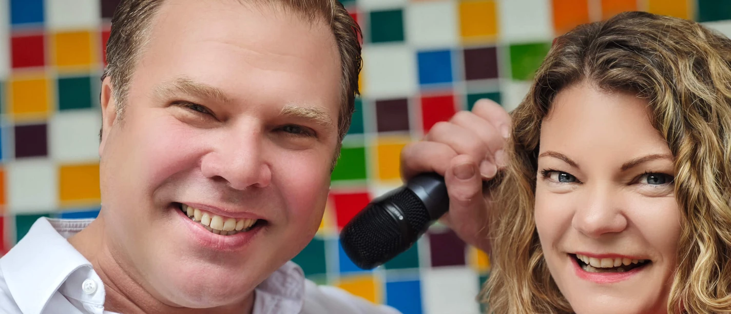 Schmitzebud Duo A smiling Schmitzebud duo with a microphone in front of a colorful tiled wall