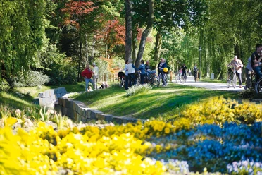 Wallanlagen Rietberg Personen genießen einen sonnigen Tag auf einem Grünstreifen mit bunten Blumen und umgeben von Bäumen.
