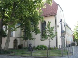 Ein Kirchengarten mit Statue und schmiedeeisernem Zaun, flankiert von altehrwürdigen Bäumen.