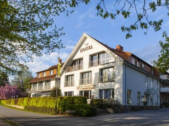 Hotel in einem großen, weißen Gebäude mit Schrägdach, umgeben von Bäumen und blühenden Sträuchern.