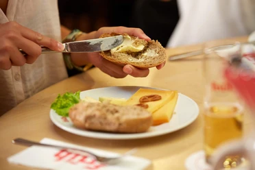 Halver Hahn Hände bestreichen ein Röggelchen mit Butter, daneben Käse und ein Glas Kölsch.