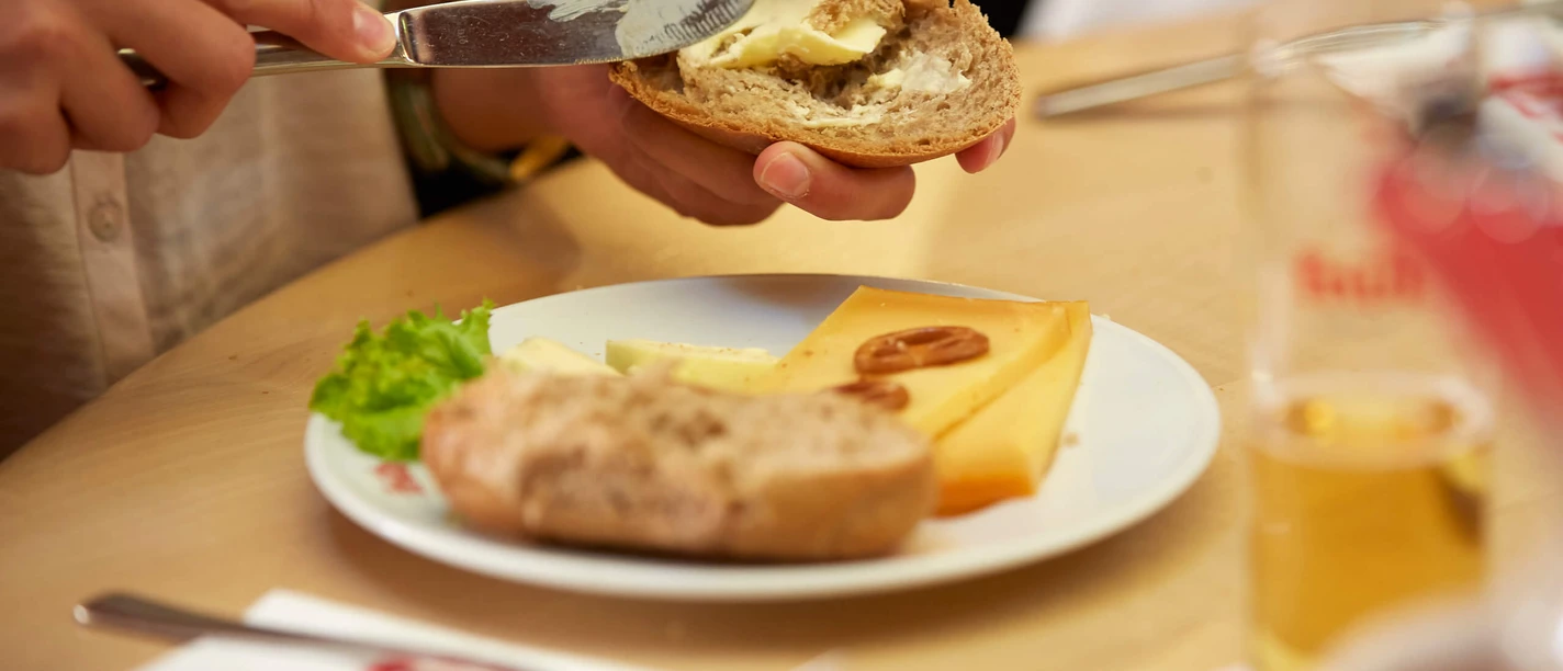 Halver Hahn Hände bestreichen ein Röggelchen mit Butter, daneben Käse und ein Glas Kölsch.