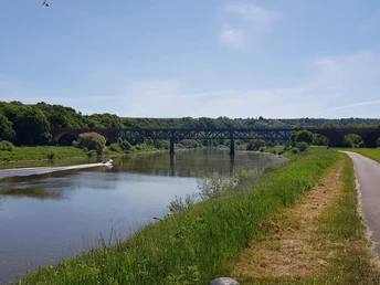 Radfahrer auf Feldweg entlang eines ruhigen Flusses mit Böschung, Brücke und Boot im Hintergrund.