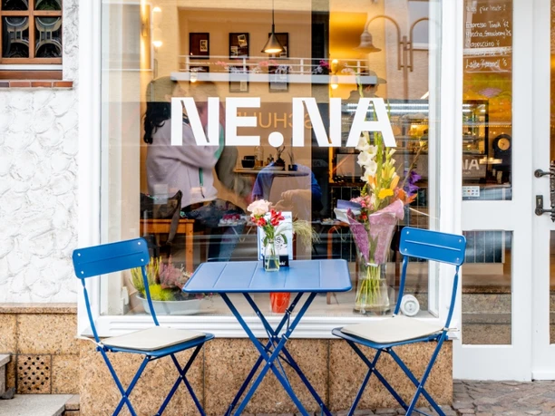 Blauer Tisch vor dem Café Ne.Na Außenansicht des NE.NA Cafés mit blauem Tisch, Stühlen und Blumen vor dem Schaufenster