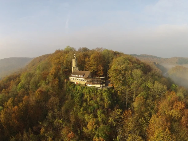 Historische Burganlage auf bewaldetem Hügel mit Aussichtsturm, von Herbstwald umgeben.
