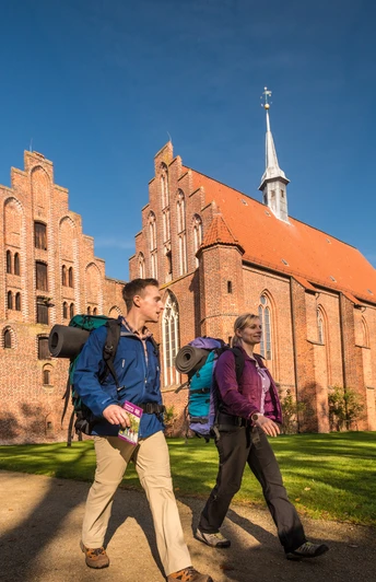 wienhausen_herbst_dk_l-neburger-heide-2015-378-l-neburger-heide-gmbh Zwei Wanderer mit Rucksäcken spazieren entlang einer beeindruckenden Backsteinkirche im Sonnenlicht.
